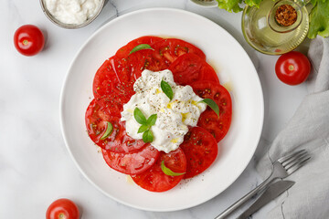 Stracciatella cheese salad with tomatoes, oil and basil on white plate on marble background. Italian Stretched curd cheese product Stracciatella.