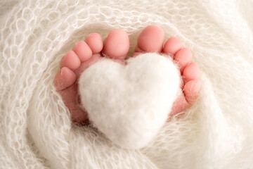 Knitted white heart in the legs of a baby. Soft feet of a new born in a white wool blanket. Close-up of toes, heels and feet of a newborn. Macro photography the tiny foot of a newborn baby. 