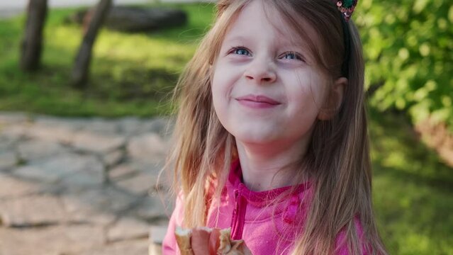 Portrait cute happy little girl with long blonde hair eating hot dog on park, smile child in pink jacket eats fast food on sunny afternoon.