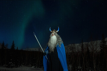 old man Odin with a staff stands in the snow at night against the backdrop of the northern lights - obrazy, fototapety, plakaty