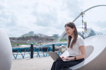 A young woman works at her laptop, sitting on a modern bench on the Newcastle waterfront, against the backdrop of Sage Gateshead and Gateshead Millennium. North East England.
