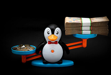Bowl of children's scales in the form of a toy penguin with a stack of paper money and metal coins on a black background