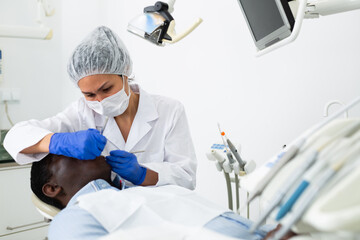 Professional asian female dentist performing teeth treatment to adult african american man in dental clinic..