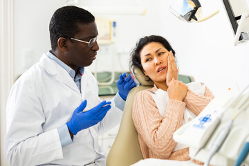 Upset Asian woman sitting in dental chair, complaining to qualified African American dentist about toothache..