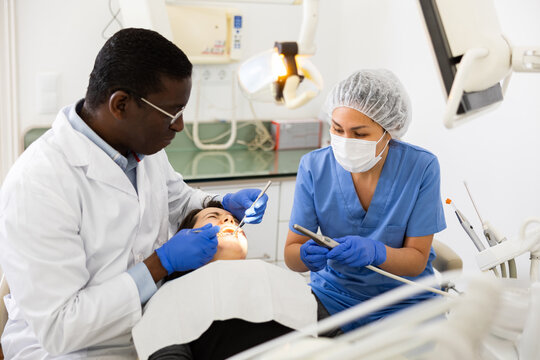 Focused african american stomatologist with female assistant treating teeth to woman patient in modern dental office..