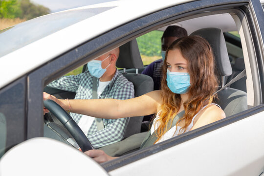 Young Woman In Protective Face Mask Driving Car With Two Male Fellow Travelers In Passenger Seats. Concept Of Individual Precautions During Covid 19 Pandemic