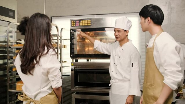 Senior Asian male chef of culinary learning course teaches and explains about electric oven to cooking class students for baking pastry dough for bread and bakery foods in stainless steel kitchen.