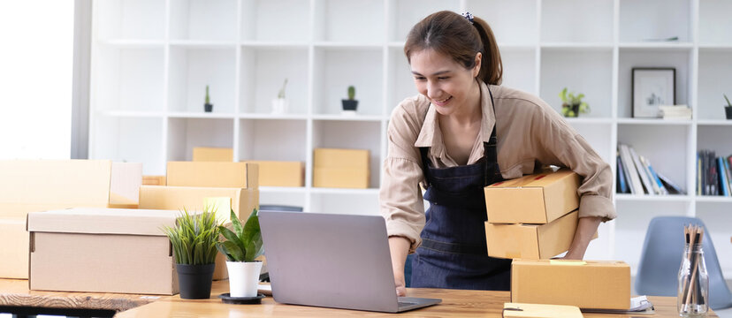 Young smiling beautiful owner asian woman freelancer sme business online shopping working on laptop computer with parcel box at home - SME business online and delivery concept