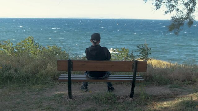 Young man sitting alone on bench overlooking rough sea