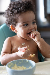 Cute Male Toddler with curly brown hair smiling while eating his breakfast in high chair and making a big mess with porridge, spoon and bowl.