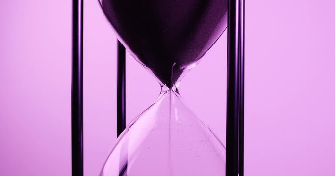 Closeup of sand timer clock with sand flowing from through hourglass on purple tone.

