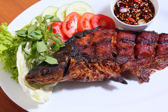 Patin fish (siamese shark, sutchi catfish), which is grilled and grilled whole. With a complement of lettuce, cabbage, basil and tomatoes. Plus spicy soy sauce. White plate. Wooden table.
