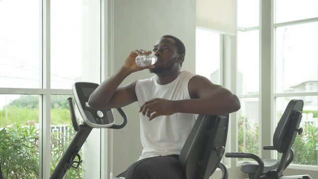Portrait of strong black african american man drinking a bottle of water, doing exercise, working out, and training in gym or fitness center in sport club. Recreation. Lifestyle activity people.
