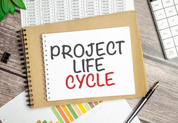 project life cycle words on notebook with pen, charts and clips on wooden background