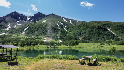 Fototapeta na wymiar On the shore of a calm alpine lake there are places to relax, canopies, benches. A mountain range against the blue sky. Reflection in the water. Kamchatka. Vachkazhets. Lake Tahkoloch