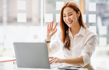 Happy young asian business woman waving hands to greeting partner during making video conference with her team.