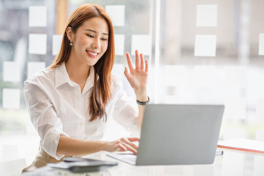 Happy young asian business woman waving hands to greeting partner during making video conference with her team.
