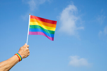 man's hand wears LGBT rainbow wristband is waving LGBT rainbow flag on background blue sky, concept for LGBTQ+ equality movement community