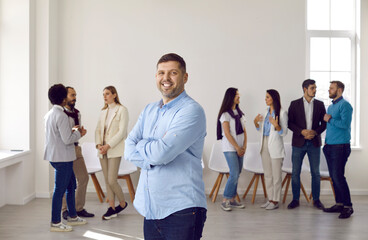 Portrait of happy male leader or boss stand forefront diverse international employees in office. Smiling young Caucasian businessman or CEO show leadership, colleagues in background.