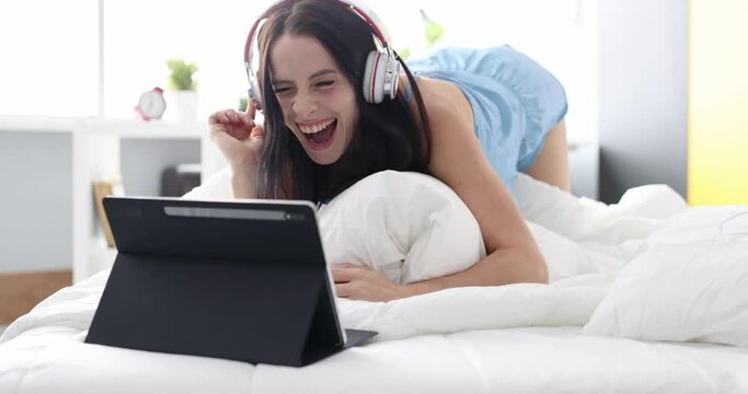 Happy woman in headphones listening to music from tablet and dancing at home in bed