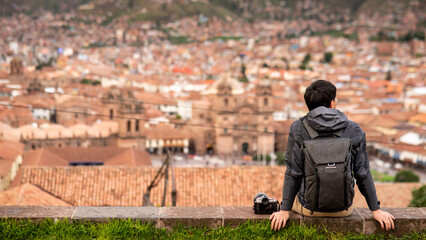Asian man tourist and photograpaher sitting on viewpoint looking at cusco city. Cusco (Cuzco) is a city in southeastern Peru, near the Urubamba Valley of the Andes mountain range - obrazy, fototapety, plakaty
