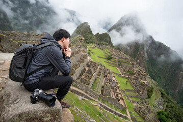Asian man tourist and photographer looking at Machu Picchu, one of seven wonders and famous tourist attraction in Cusco Region of Peru. This majestic place has known as Lost City of the Incas. - obrazy, fototapety, plakaty