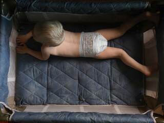 A three-year-old child in pampers diapers is lying in a crib. Naked kid plays and indulges in a small bed in the arena - obrazy, fototapety, plakaty