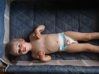 A three-year-old child in pampers diapers is lying in a crib. Naked kid plays and indulges in a small bed in the arena