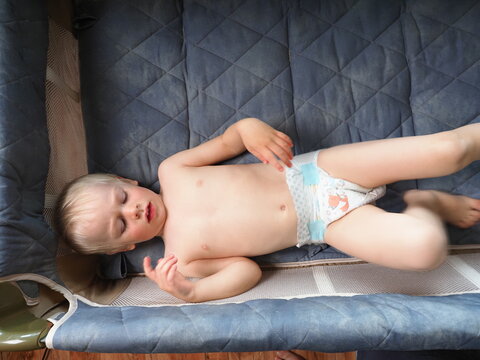 A three-year-old child in pampers diapers is lying in a crib. Naked kid plays and indulges in a small bed in the arena