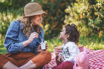 A young single mother spoon feeds yogurt to her little daughter sitting on the grass during a picnic outdoors. Concept of family, outdoor life and separated parents. - Powered by Adobe