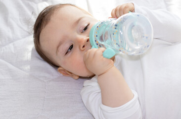 adorable baby boy laying on bed drinking water from plastic bottle with silicone head for mouth. toddler hold legs up. cute baby feet.cup with 2 handles,anti leak, straw wide mouth.  