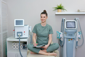 Cosmetologist is standing at laser hair removal machine with folder for recording clients and smiling. Woman in green coat doctor of beauty salon waiting for client