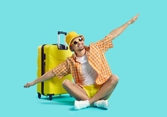 Happy man who goes on summer vacation by air poses with suitcase on light blue background. Guy in summer clothes sits with outstretched arms imitating flight of airplane. Air flight journey concept.