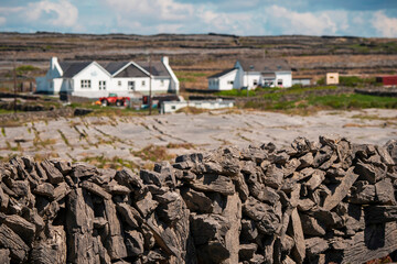 Rough stone fence in focus. White houses and white cloudy sky out focus. Rough stone terrain of...