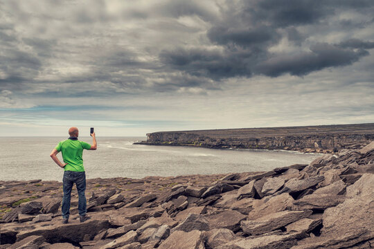 Male tourist on a rough stone surface of a cliff taking picture on phone. Ocean and dramatic cloudy sky in the background. Aran island, Ireland. Stunning Irish landscape. Travel and tourism concept.