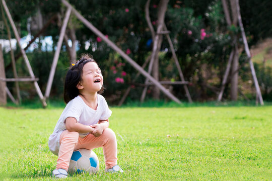 Portrait image of child 4-5 years old. Cute Asian girl is resting on soccer ball after having fun running and kicking it. Happy children smile and laugh cheerfully. During summer or spring time.