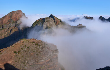 Panorama from Pico do Areeiro, a starting point of PR1 hiking trail in the Madeira Island. Hikers on the trail to the Pico Ruivo. Fog ascending from a valley and remaining among mountain folds.