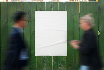 Poster mockup on a green wooden fence. People pass by the poster. Blank poster for design presentation