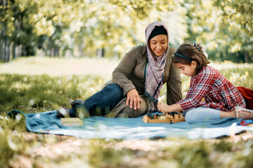 Happy Muslim mother plays chess with her daughter in park.