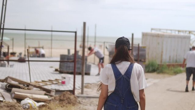 The female worker in overalls and cap goes on the construction site. Woman with long brune haire coming near a building under construction by the sea.