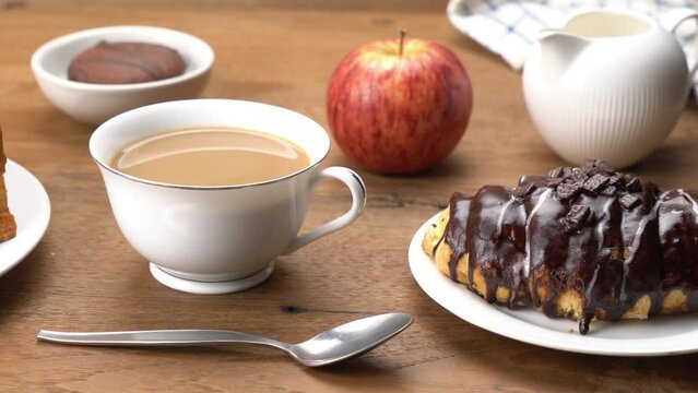Moving onthe breakfast table with a chocolate covered croissant, a cup of milk coffee, a loaf of bread, a piece of choco pie and a ripe red apple on wooden table.