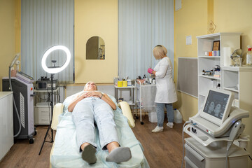 Beautician prepares to perform cosmetic procedures while patient lies on table for beauty...