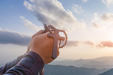 Hands folded holding rosary crucifix symbol with bright sunbeam on the clouds sky background.Religion and Hope Concept. - obrazy, fototapety, plakaty