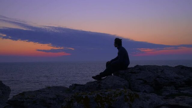 Young man at sea shore waits for the sunrise, gazing at the horizon, sitting on a rock. General view