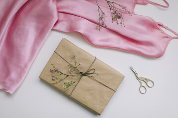 Composition with pink silk fabric, gift in craft paper and threads and scissors on white background. Flat lay, top view. Hobby, leisure concept.