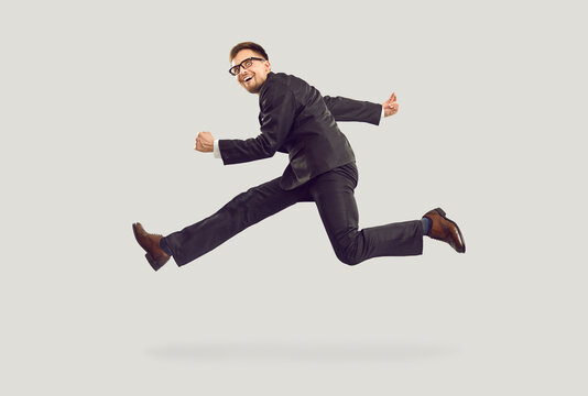 Crazy excited young businessman in black suit running and jumping isolated on gray background. Side view of man who seems to be frozen in air with crazy expression seeks to achieve career and wealth.