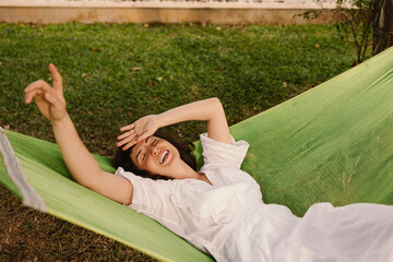 Funny young caucasian girl wear dress smiles and squints from sun lying in hammock summer. Brunette wears white spacious sundress in warm weather. Summer playful mood concept - obrazy, fototapety, plakaty