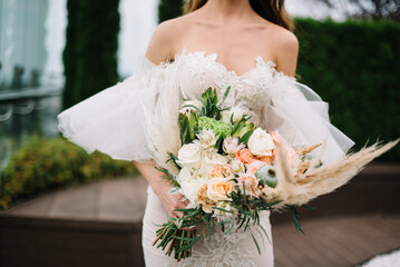 stylish bride in a dress standing in a green garden and holding a wedding bouquet of flowers and greenery