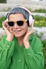 Cheerful senior woman listening to music in park. Female model with short grey hair in bright clothes and big headphones. Leisure, activity, technology concept