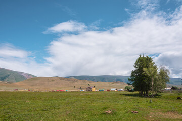 Obraz premium Dramatic alpine view from trees to small houses in sunlit green meadow against high mountain range under clouds in blue sky. Scenic mountain landscape at changeable weather. Village in high mountains.